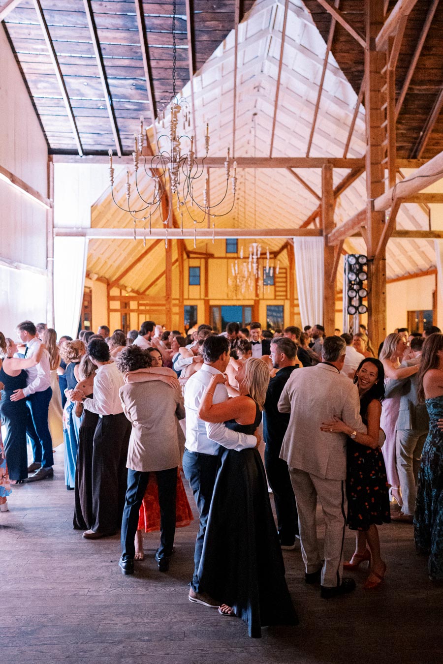 A lively wedding reception in a rustic barn setting with guests dancing under elegant chandeliers, creating a warm and joyful atmosphere.