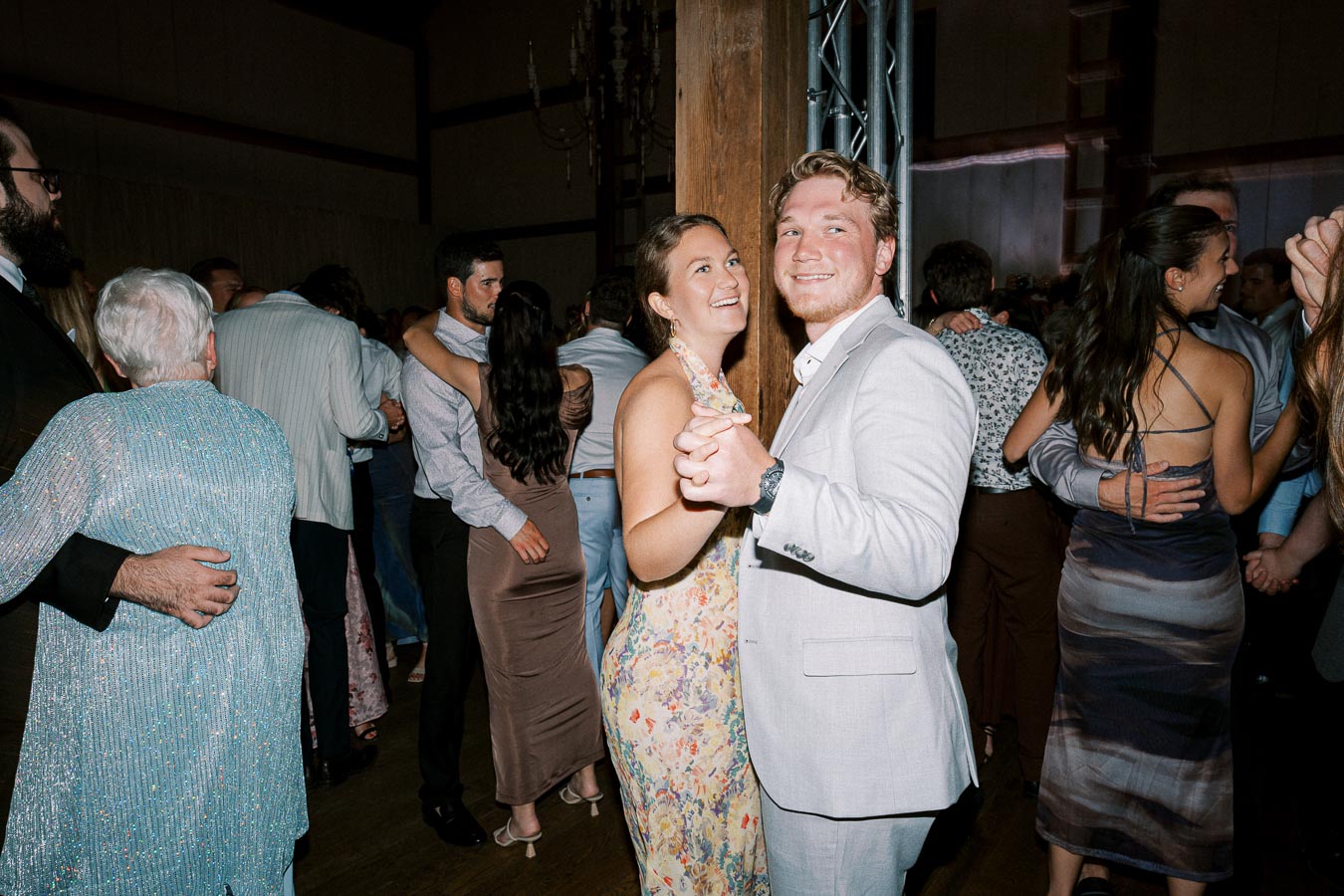 Group of people dancing and enjoying themselves at an indoor event or celebration, with a couple in the foreground smiling at the camera.