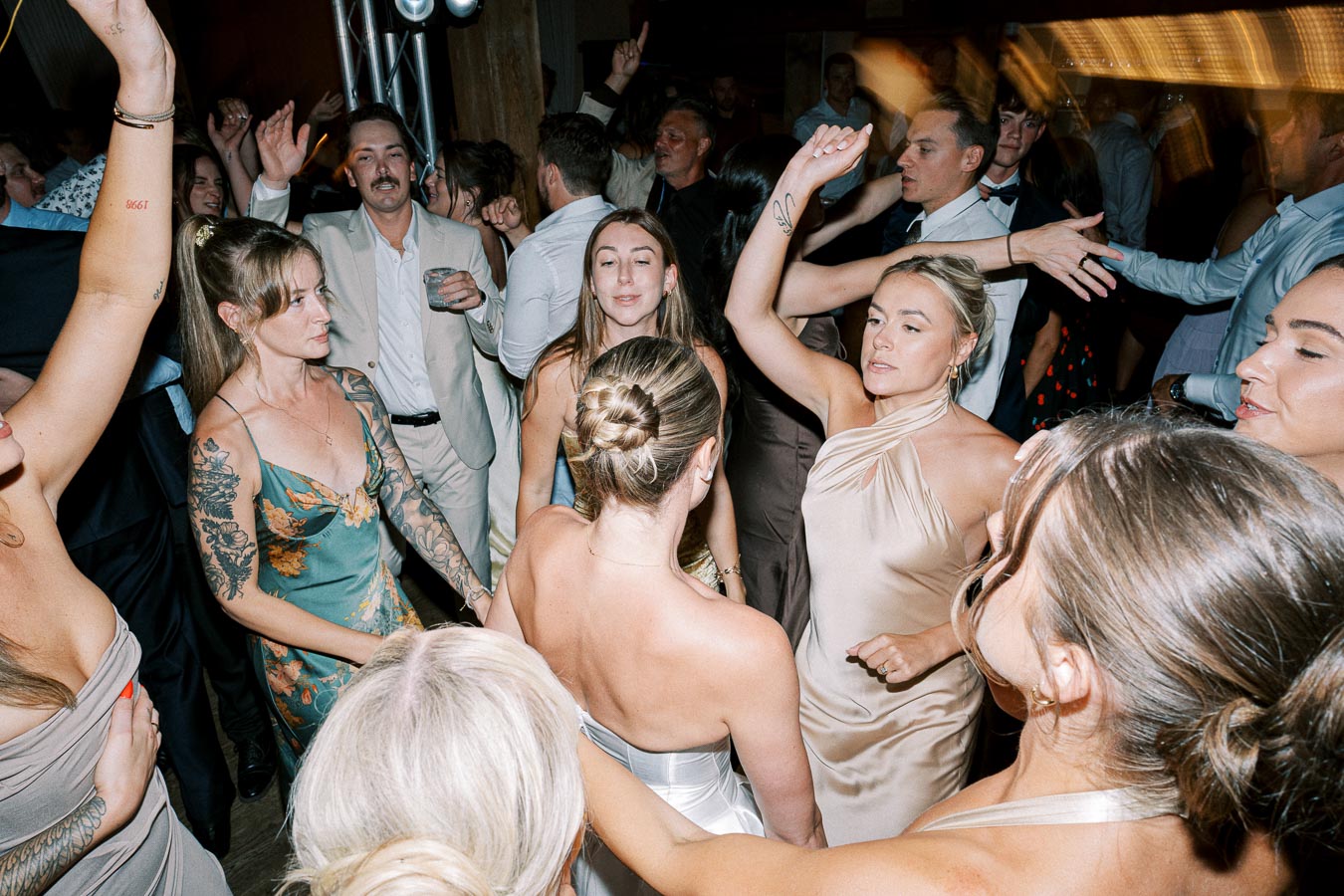 A lively group of people dancing and mingling at a party, with women in elegant dresses and men in suits. The room is filled with festive energy as guests enjoy the celebration.