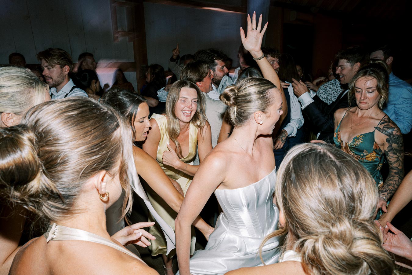 Group of people joyfully dancing at a wedding reception, with women in elegant dresses and men in formal attire celebrating together on the dance floor.