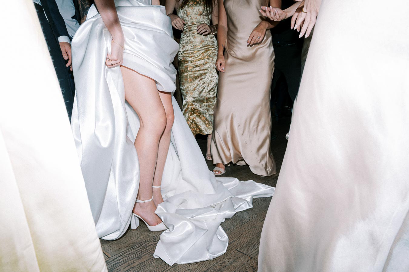 Elegant wedding reception dance floor scene with people wearing stylish dresses and satin gowns, highlighting a flowing white wedding dress and high heels on a wooden floor.