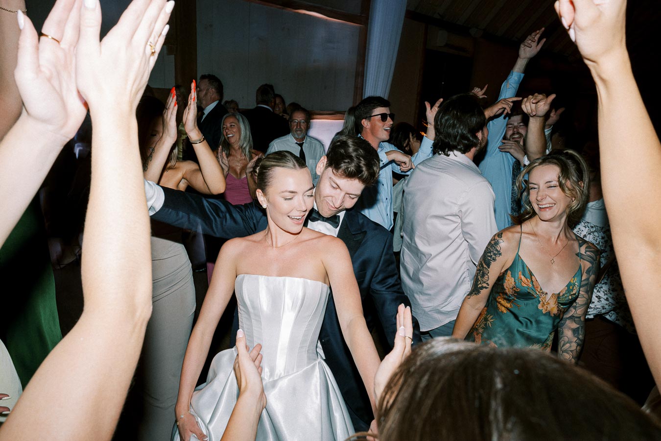 A joyful wedding celebration with guests dancing; a smiling couple in formal attire is at the center, surrounded by a lively crowd.
