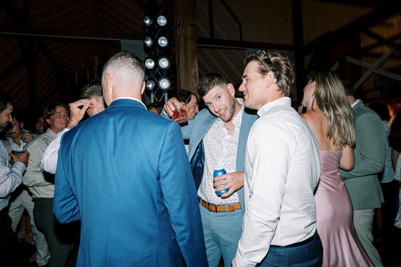 Group of people enjoying a lively party, dressed in formal attire, with one person holding up a drink and others smiling and interacting in a dimly lit venue.