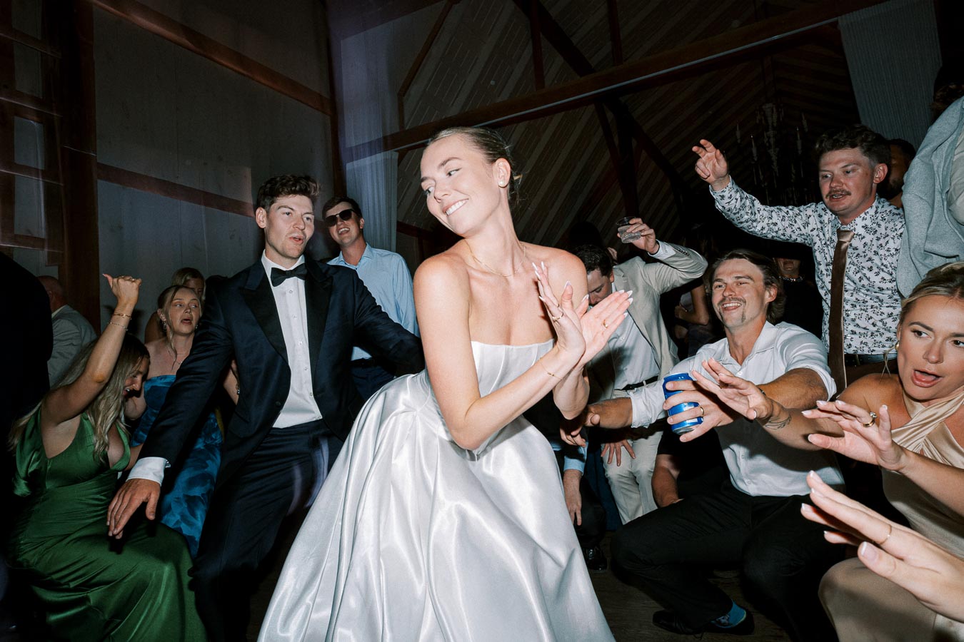 Group of people joyfully dancing at a wedding reception, with a woman in a white dress leading the celebration, surrounded by guests in formal attire.