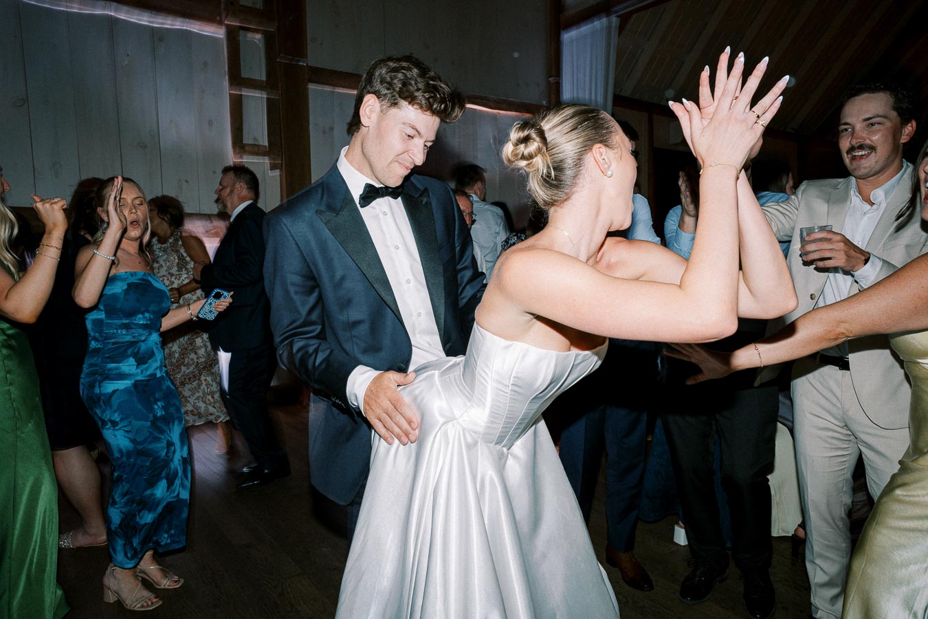 A lively wedding reception scene with guests in formal attire dancing joyfully. A man in a tuxedo and a woman in a white gown are leading the celebration amidst a group of smiling attendees.