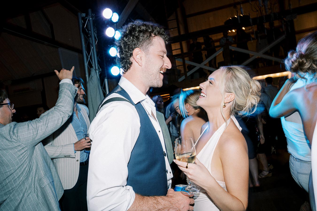 A smiling couple dancing and enjoying a celebration at a lively party with colorful lights and people socializing in the background.