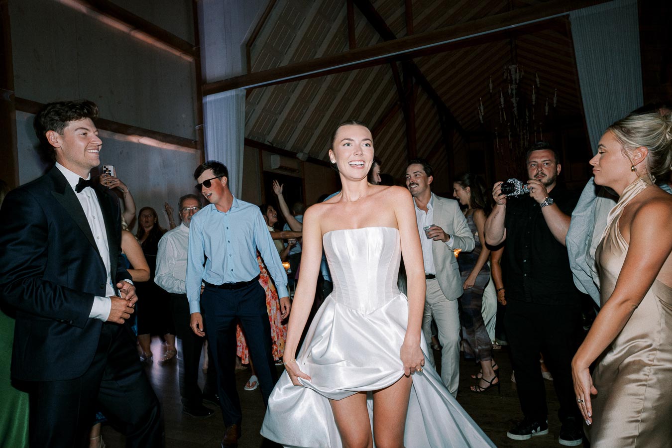 A joyful wedding celebration with guests dancing in elegant attire at a nighttime reception, featuring a bride in a stunning, strapless white gown amid a lively crowd.