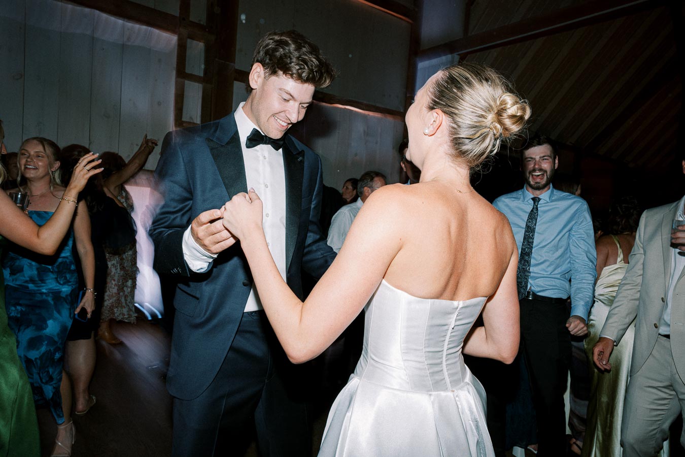 A couple elegantly dances at a wedding reception, with the bride in a strapless white gown and the groom in a black tuxedo.