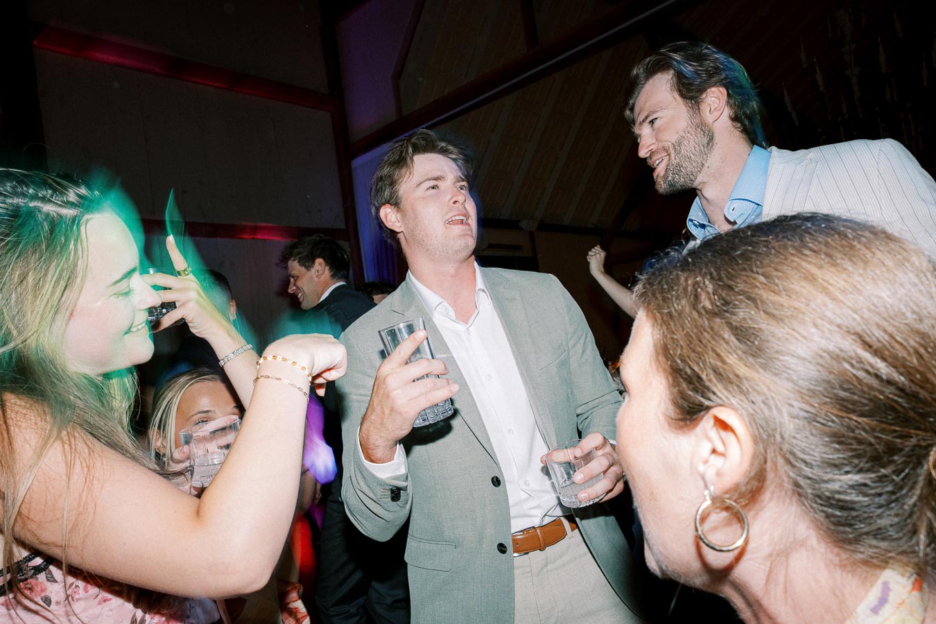 A group of people enjoying a lively party, with a man in a light suit holding drinks and engaging in conversation amidst colorful lighting and a vibrant atmosphere.