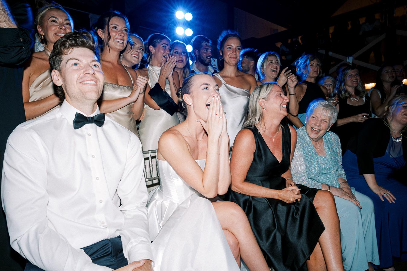 A group of elegantly dressed people, including women in satin dresses and men in formal attire, sitting and standing, excitedly watching an event with joyful expressions. The setting appears to be a lively celebration or party.