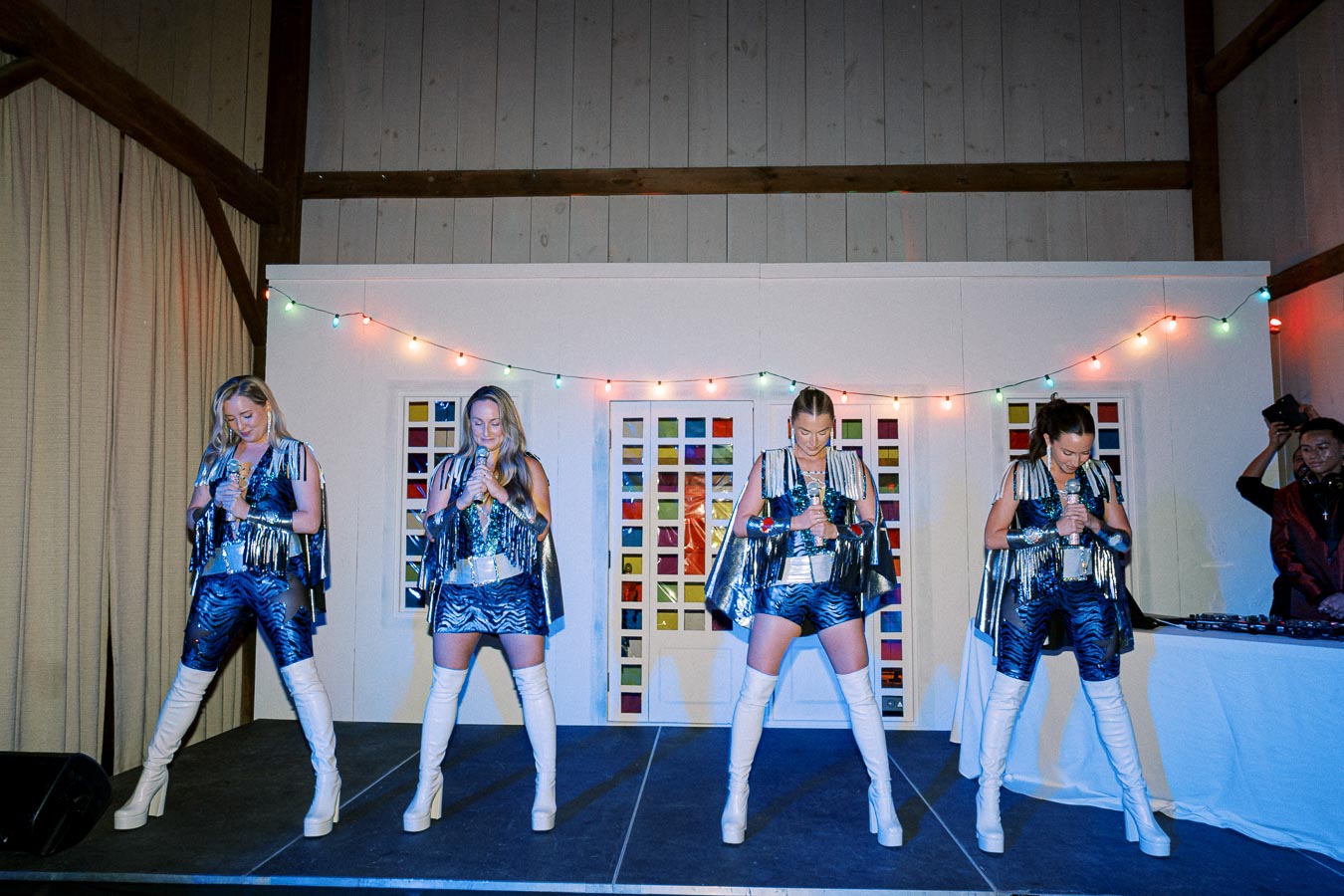 Four performers in matching blue sequined outfits and white knee-high boots sing on a stage decorated with colorful string lights. The background features white panels with colorful square accents, creating a festive ambiance.