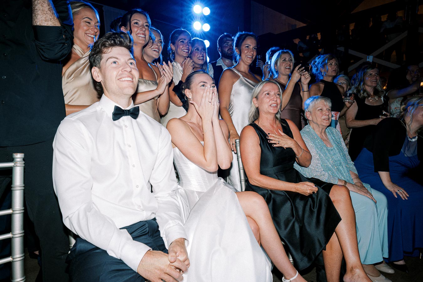 Bridal party and guests joyfully celebrating at a wedding reception, with the bride in a white gown and groom in a bow tie, surrounded by smiling family and friends.