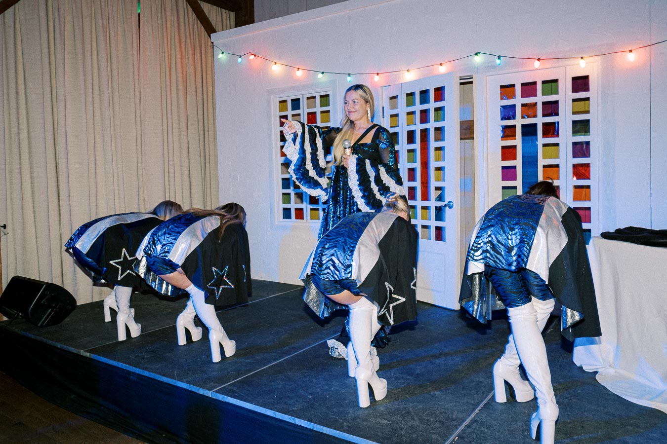 Performer on stage in a colorful costume holding a microphone, with dancers in matching outfits and high white boots bending over, against a backdrop of colorful string lights and patterned sliding doors.