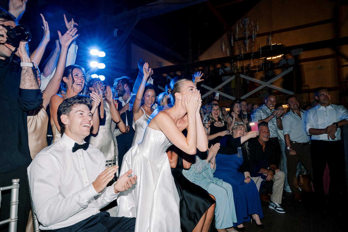A joyful wedding celebration scene with guests clapping and cheering. A bride in a white dress is excitedly covering her mouth, surrounded by happy friends and family, in a warmly lit event venue.