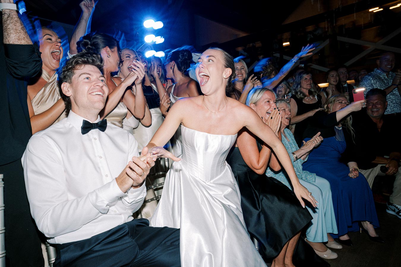 A joyous bride in a strapless wedding dress celebrates with a smiling groom in a bow tie at a lively wedding reception, surrounded by cheering and applauding guests.
