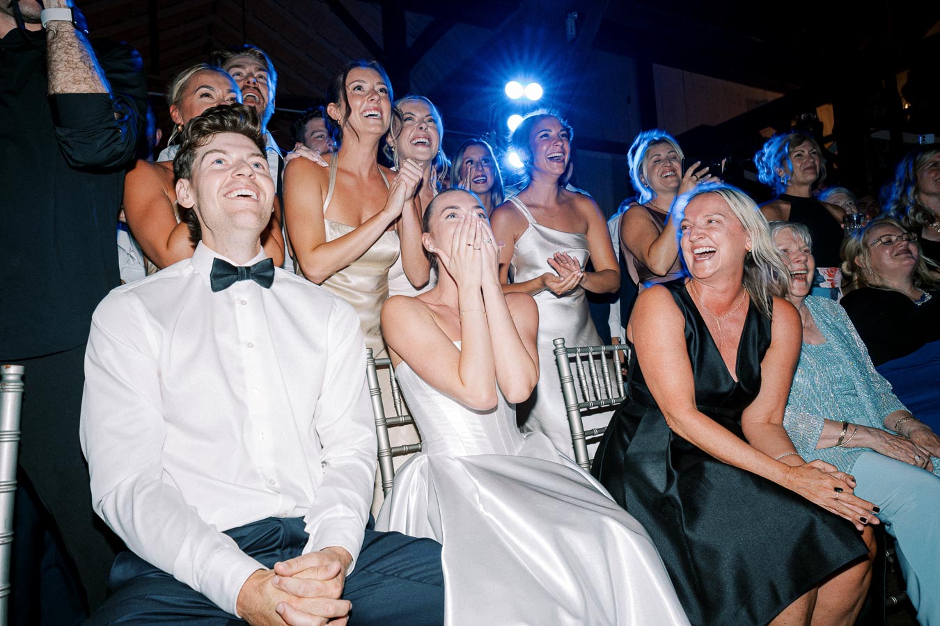 A joyful group of wedding guests in formal attire cheer and smile in excitement, sitting on silver chairs under bright blue lighting at a wedding reception.