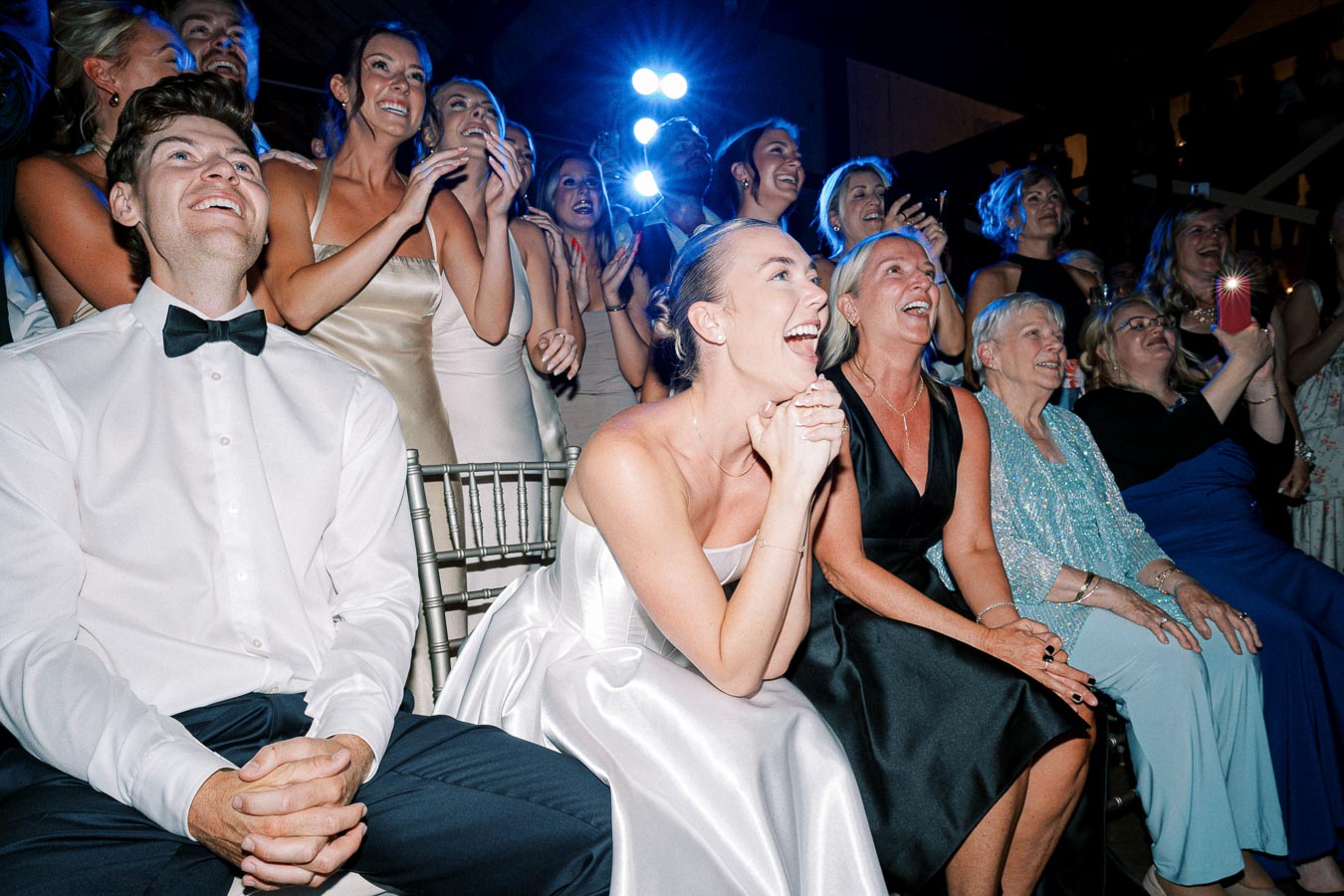 A group of well-dressed guests joyfully watching an event at a wedding reception, with women in elegant dresses and a man in a tuxedo, seated and smiling in a dimly lit venue.