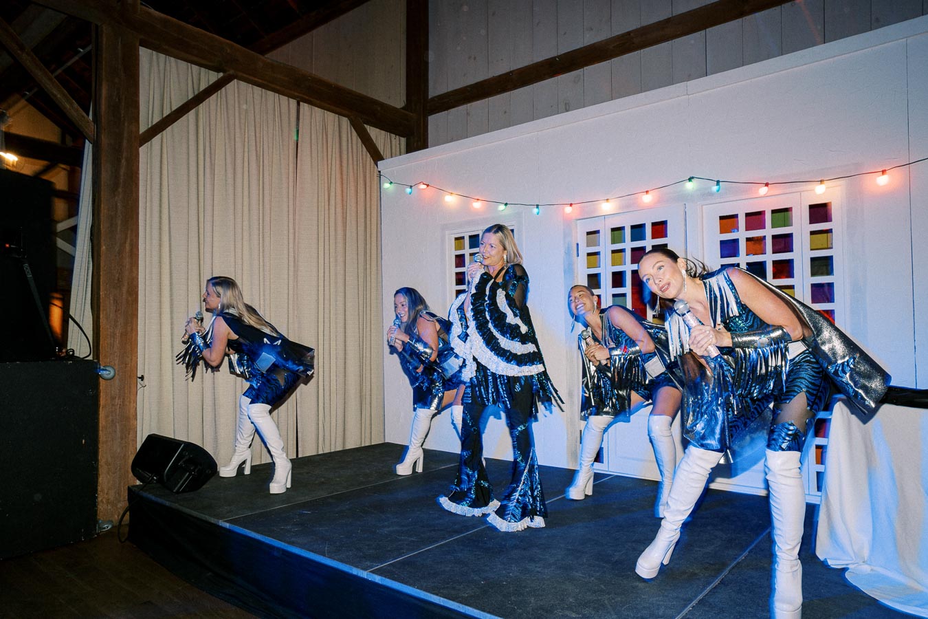 A music group performs energetically on stage in colorful costumes, holding microphones, with festive lights in the background.
