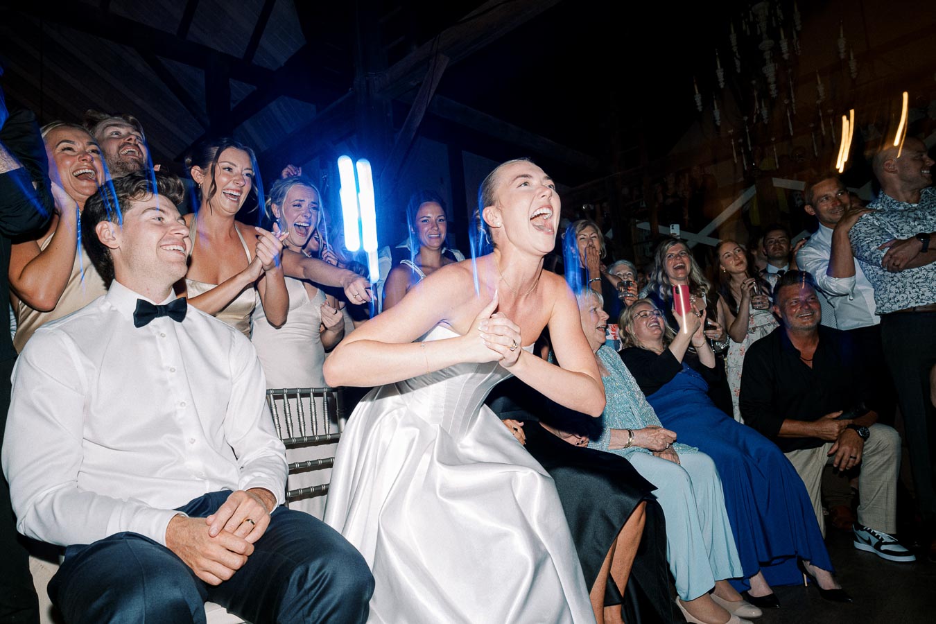 A joyous bride in a white wedding gown shares a laugh with a groom in a tuxedo, surrounded by cheering guests at a lively wedding reception.