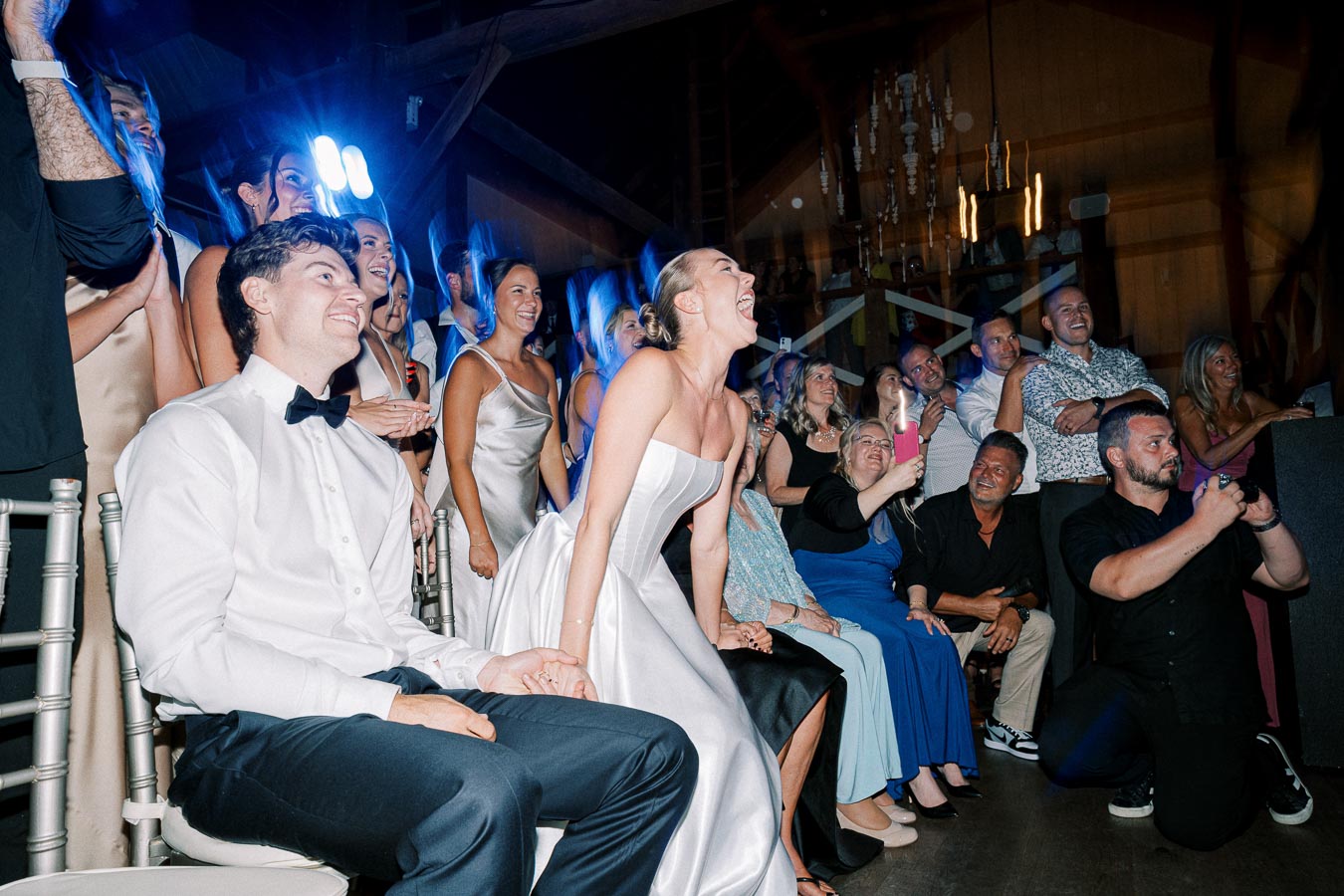 Group of people joyfully gathered at a wedding, with a bride in a white gown and a groom in a tuxedo sitting together, surrounded by friends and family celebrating in a warmly lit venue.