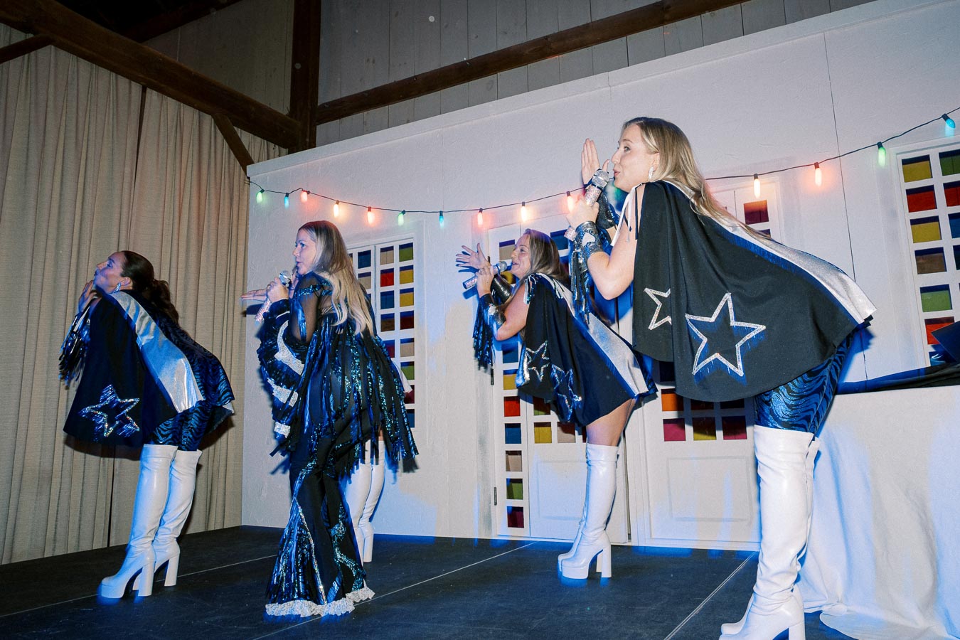 Four performers in black and silver star-themed outfits sing and dance on stage, wearing white platform boots, with colorful string lights and a patterned backdrop enhancing the vibrant atmosphere of the performance.