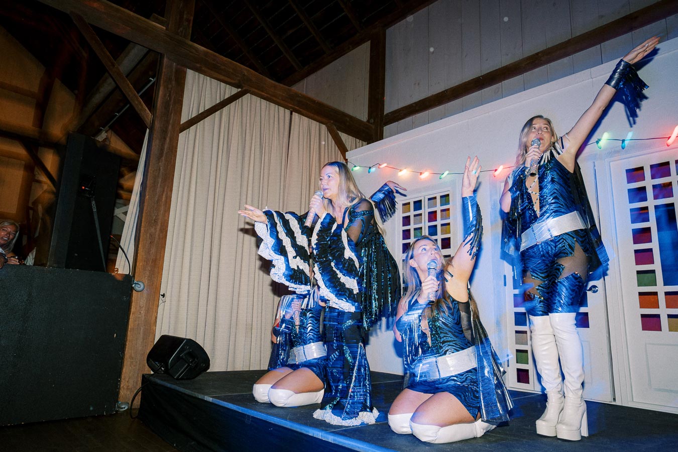 Three performers in matching blue and black costumes sing and pose energetically on stage at a lively event, with colorful lights in the background.