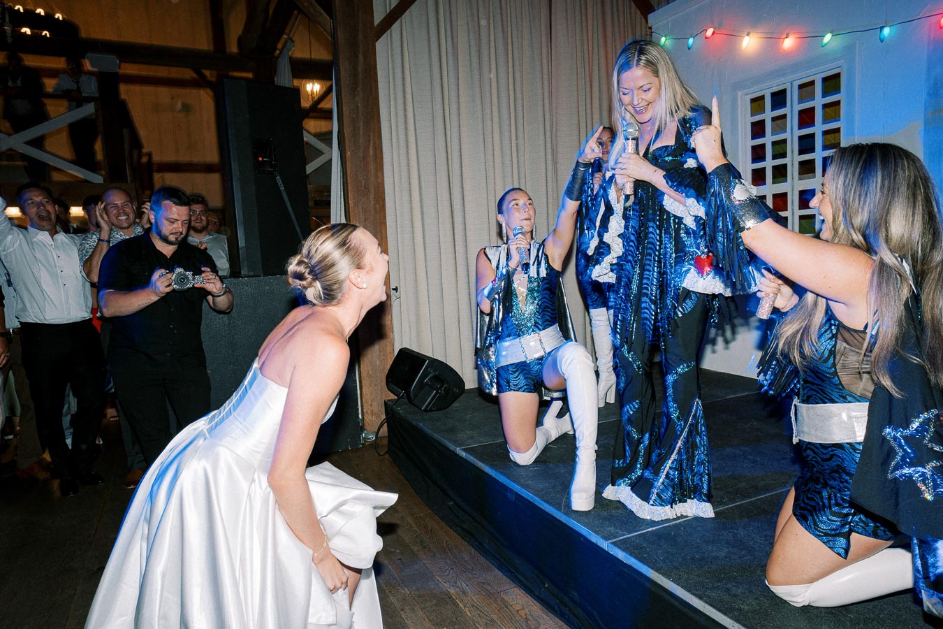 Bride laughing at a lively performance by women in blue sequined costumes during a wedding celebration, with an enthusiastic crowd capturing the moment.