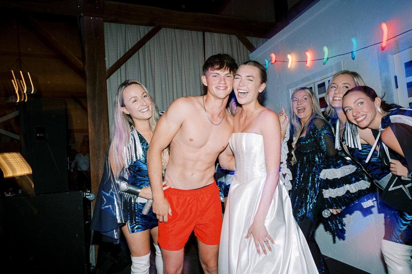 A joyful group celebration with a shirtless man in red shorts and a woman in a white dress, surrounded by friends in vibrant blue costumes, under colorful string lights at an indoor party.