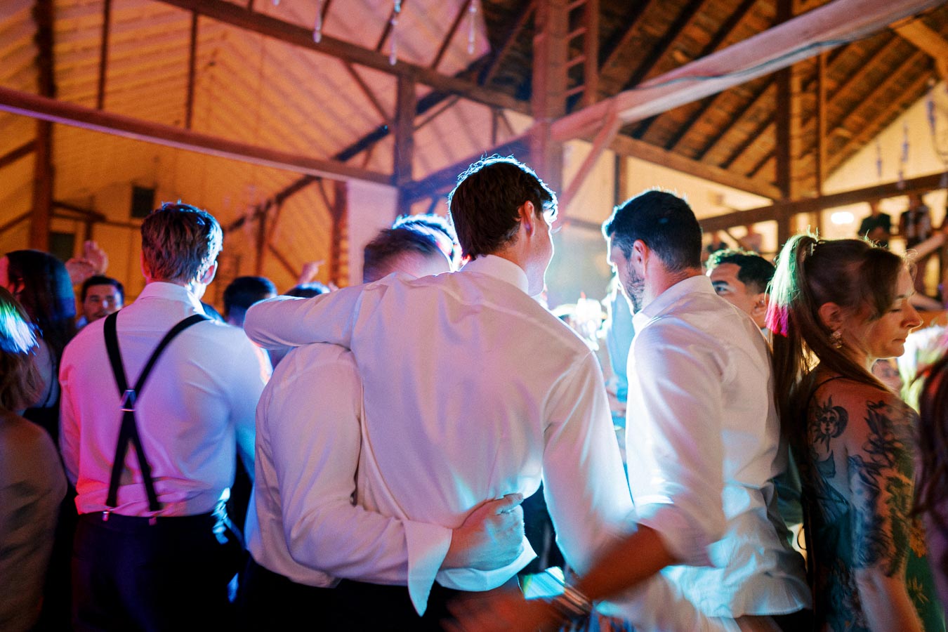 Group of people dancing and celebrating at a lively indoor party, featuring colorful lights and a warm rustic setting.