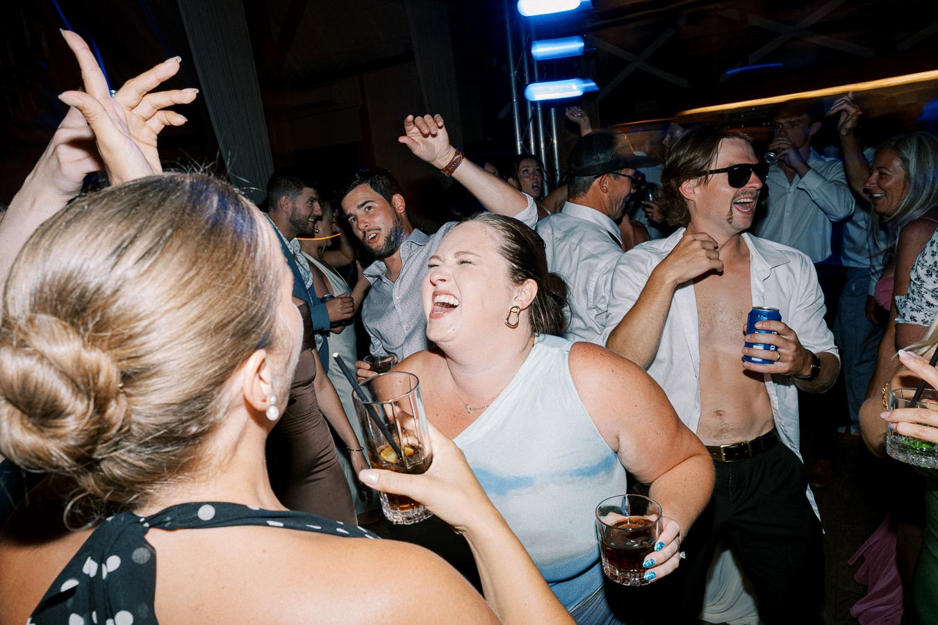 Group of people enjoying a lively party, laughing and dancing with drinks in hand, under vibrant blue lights.