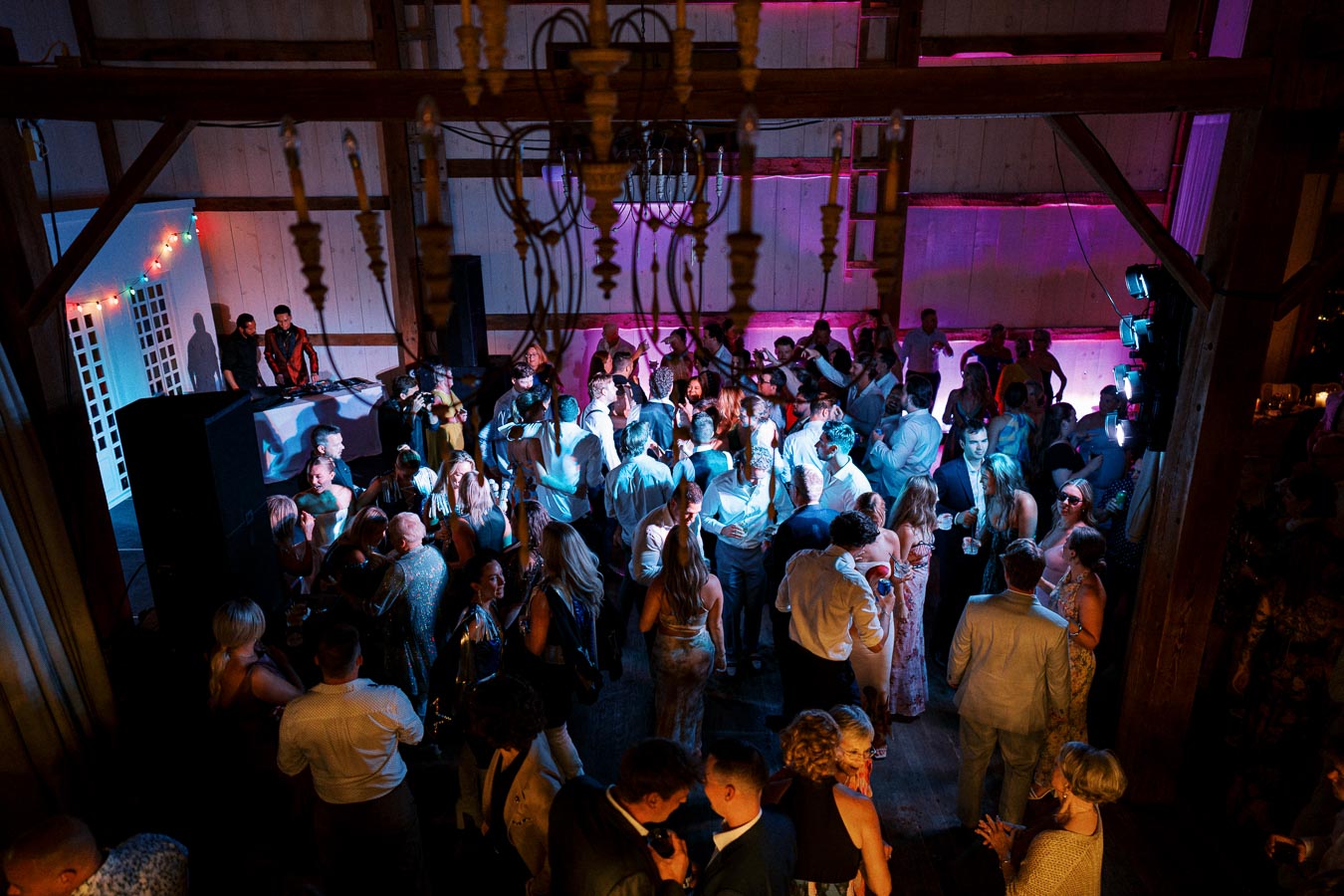 A lively group of people dancing and socializing in a dimly lit barn-themed venue with a DJ playing music and colorful lights enhancing the festive atmosphere.