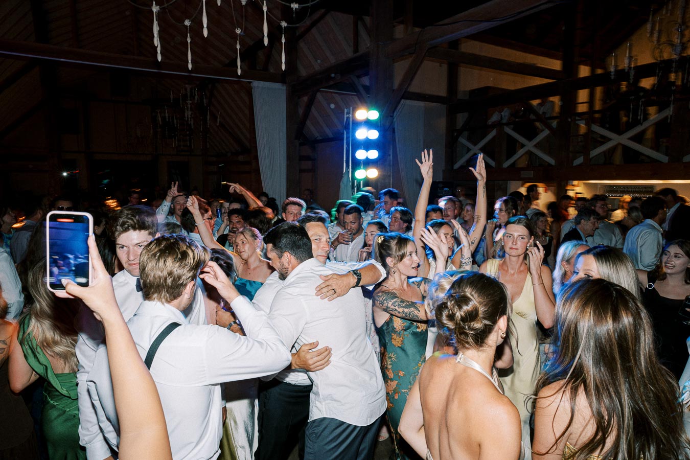 A lively wedding reception party with guests dancing energetically in a dimly lit hall, colorful lights in the background, and a mix of formal and casual attire.