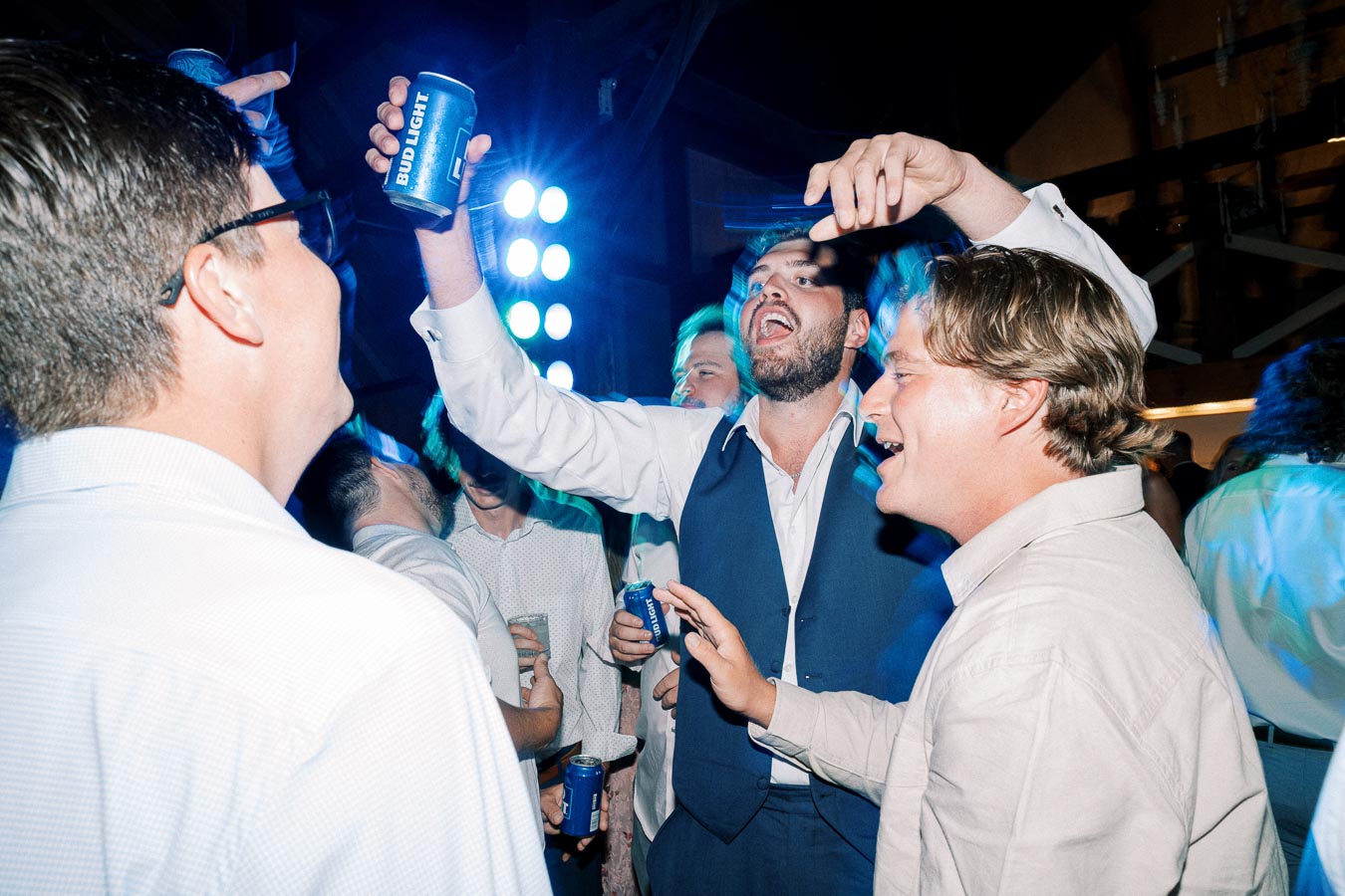 Group of people enjoying a lively party, holding Bud Light cans, under vibrant blue lighting.