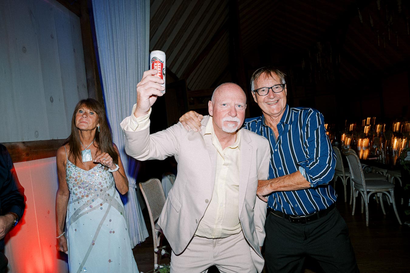 A joyful celebration scene at a lively party, featuring a man in a beige suit holding up a can of Stella Artois beer, surrounded by smiling friends in a warmly lit venue.