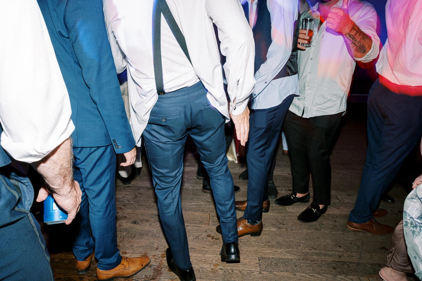 Group of well-dressed men socializing on a dance floor at a festive event, wearing suits and dress shoes, while one person gives a thumbs-up, creating a lively atmosphere.