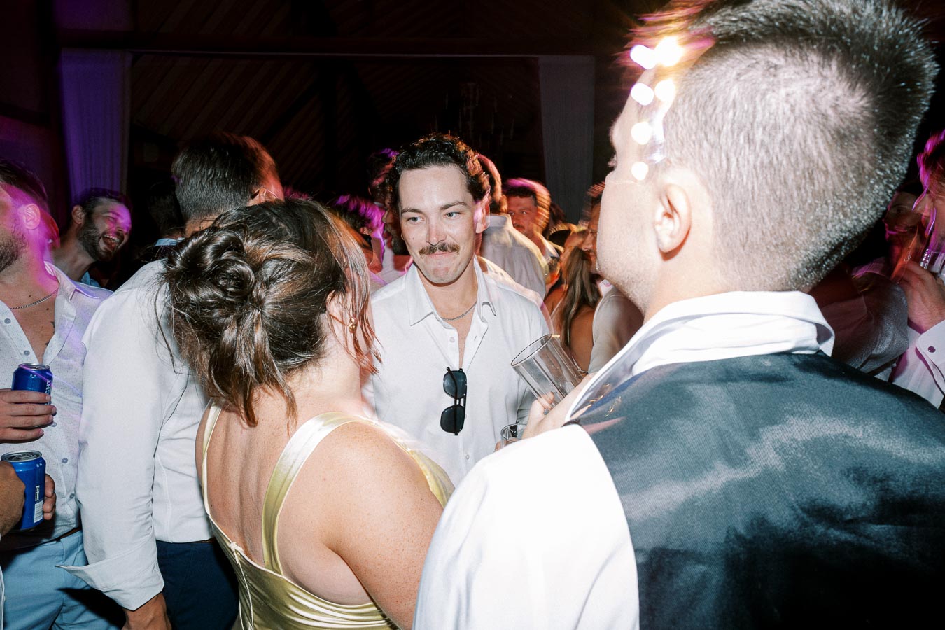 A lively party scene with a group of people socializing on a dance floor. They are dressed in formal attire, enjoying drinks and conversation under colorful lights, creating a vibrant and festive atmosphere.