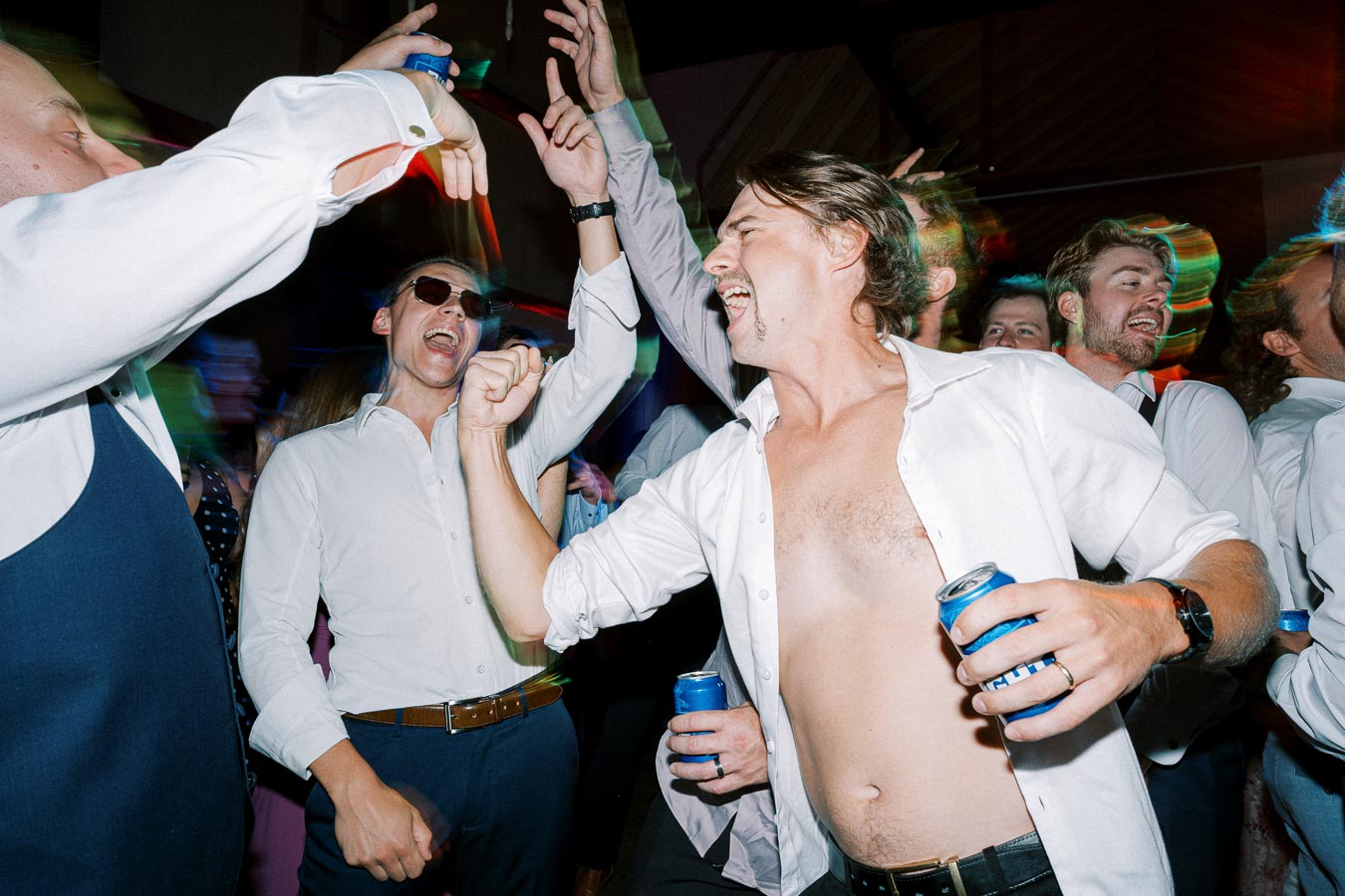 A group of men energetically dancing and enjoying a party, dressed in casual white shirts, with one holding a canned beverage. The vibrant atmosphere is highlighted by colorful lights and lively expressions, capturing the essence of a joyful celebration.
