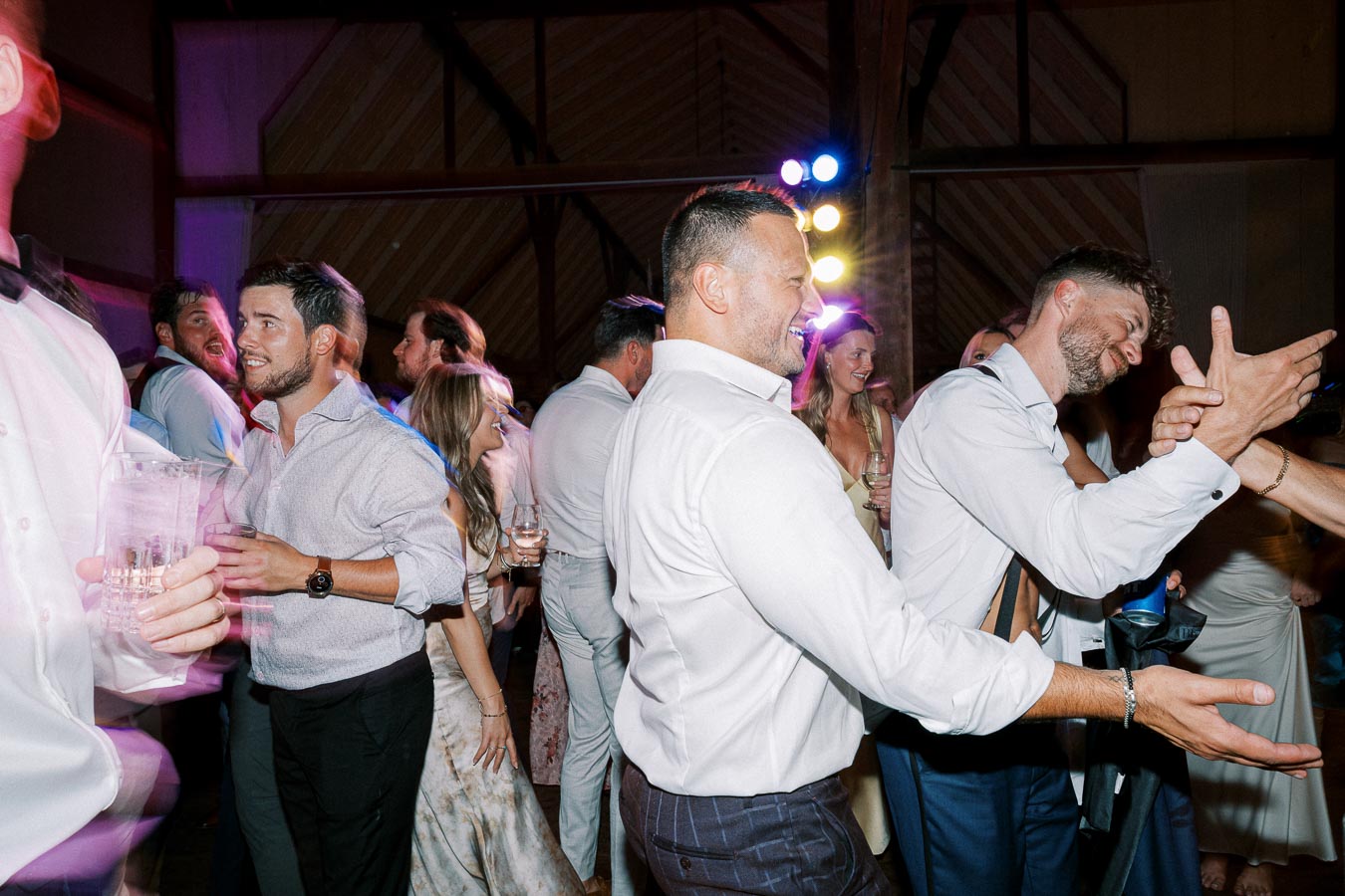 A lively group of people enjoying a celebratory event, dressed in formal attire, dancing and smiling under vibrant party lights in a wooden venue.