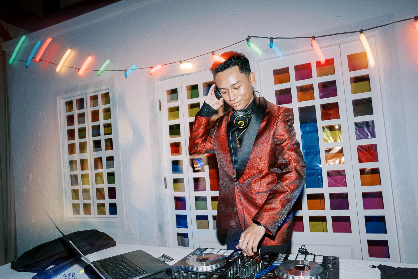 DJ in a stylish red jacket performing at an event, with colorful string lights and a laptop in the background.