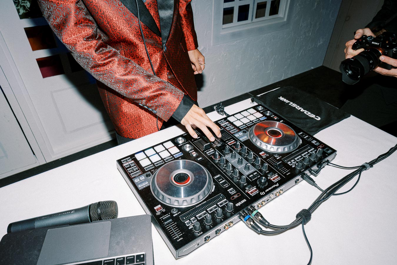 A DJ in a stylish red jacket mixing music on a professional Pioneer DJ controller at an event, with nearby equipment including a microphone and a camera.