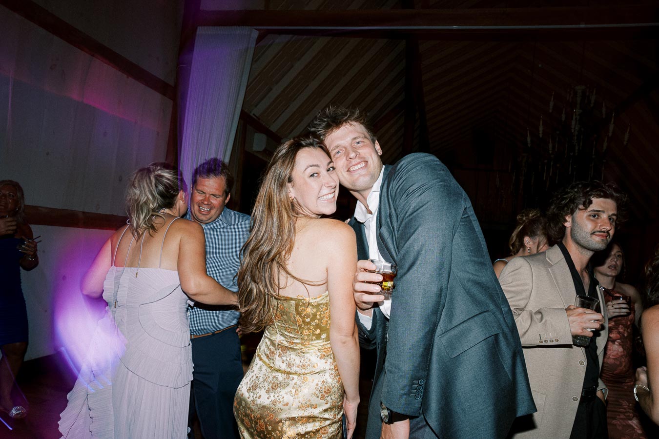 Group of elegantly dressed people enjoying a lively party, with two smiling friends in the foreground holding drinks, and others dancing and socializing in a dimly lit venue with colorful lights.