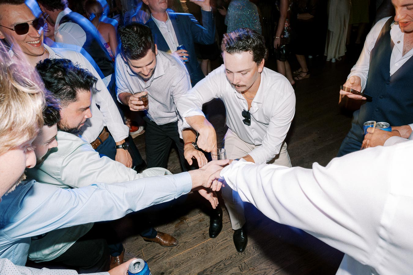 Group of men having fun and playing a party game on a dance floor, surrounded by friends and energetic lighting.