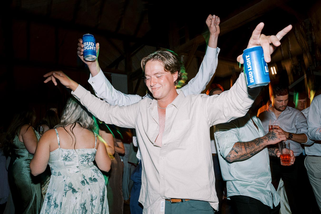 A group of people enjoying a lively party, holding drinks and dancing energetically in a dimly lit venue, with one person prominently holding a beer can and smiling with eyes closed, capturing the essence of a fun night out.