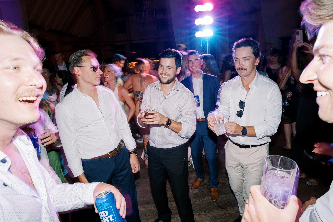 Group of people enjoying a party with drinks, dancing, and colorful lights in the background.