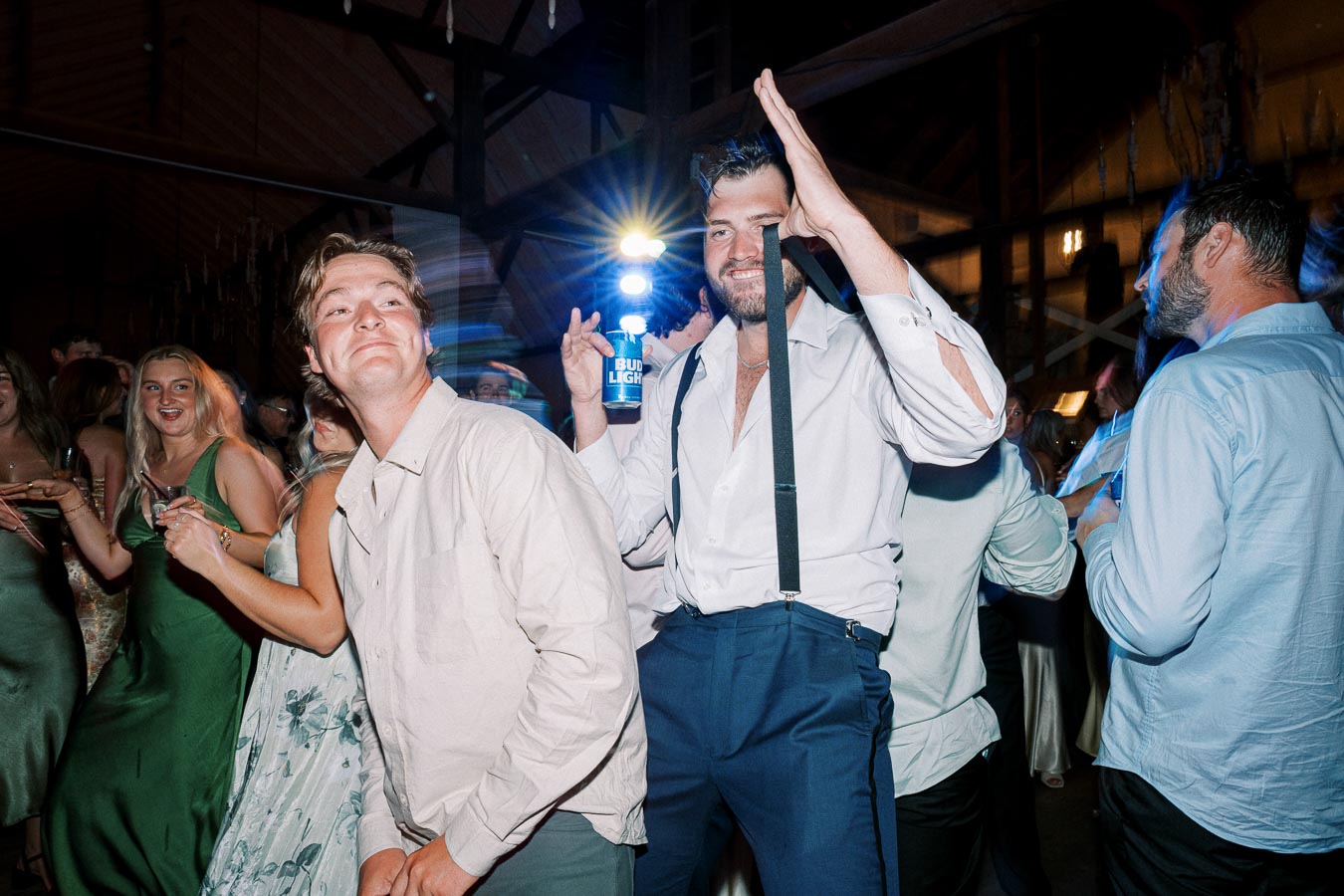 A lively wedding reception party with guests dancing and enjoying themselves on the dance floor. A man in suspenders holds a drink, surrounded by laughing friends, under colourful lights in a festive atmosphere.