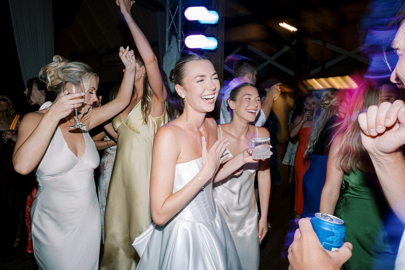 A group of people in elegant dresses enjoying a lively party, holding drinks, and dancing with colorful lights in the background.