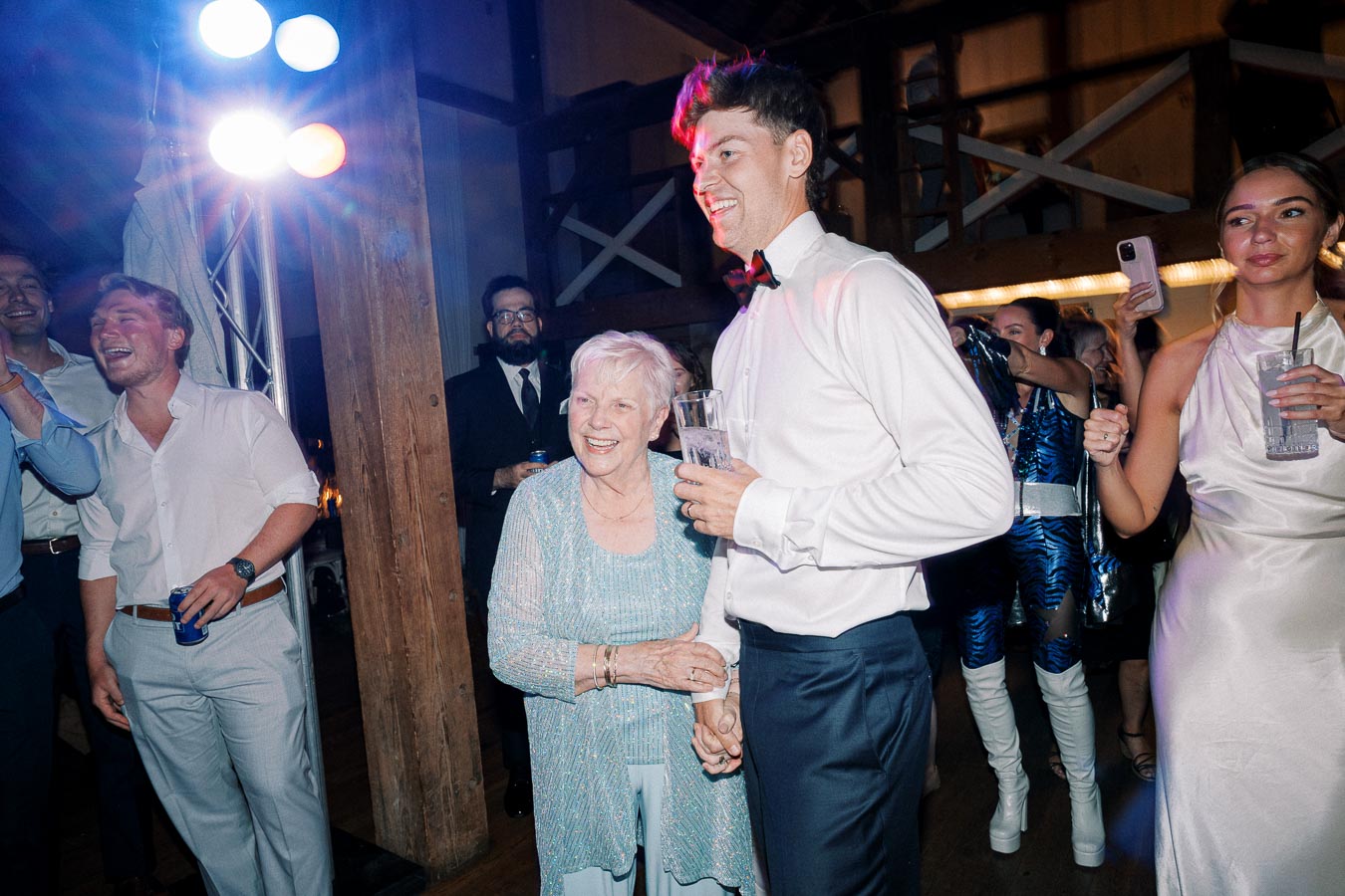 A joyful scene at a party with diverse guests dancing and smiling, including an elderly woman in a blue outfit and a young man in formal attire, highlighting multigenerational celebrations.