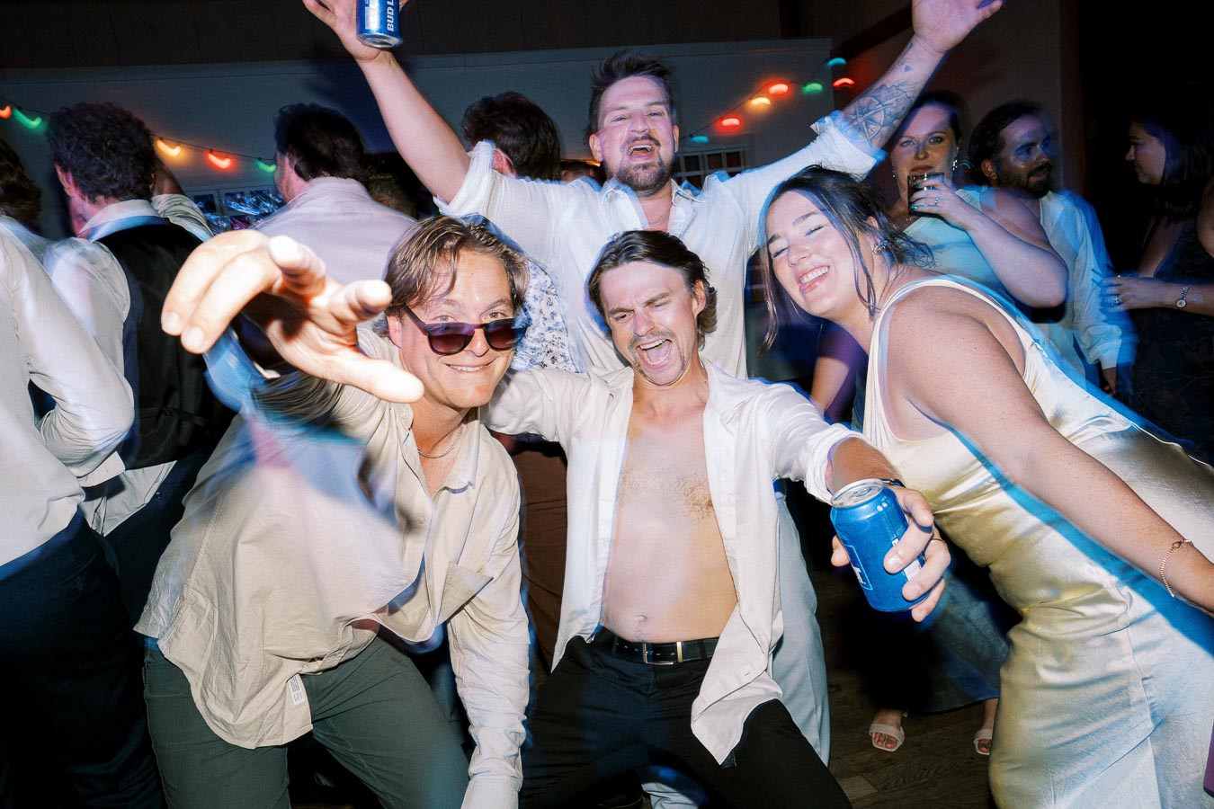 Group of friends celebrating at a lively party, holding drinks and dancing under colorful lights.