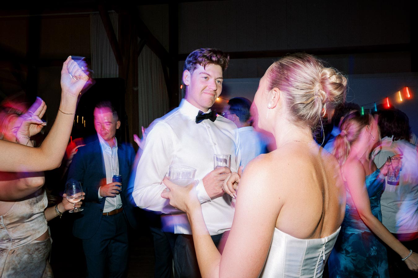 A lively wedding party scene with elegantly dressed guests socializing and dancing, featuring a man in a white shirt and bow tie engaging with a woman in a strapless dress, colorful lights adding a festive atmosphere.
