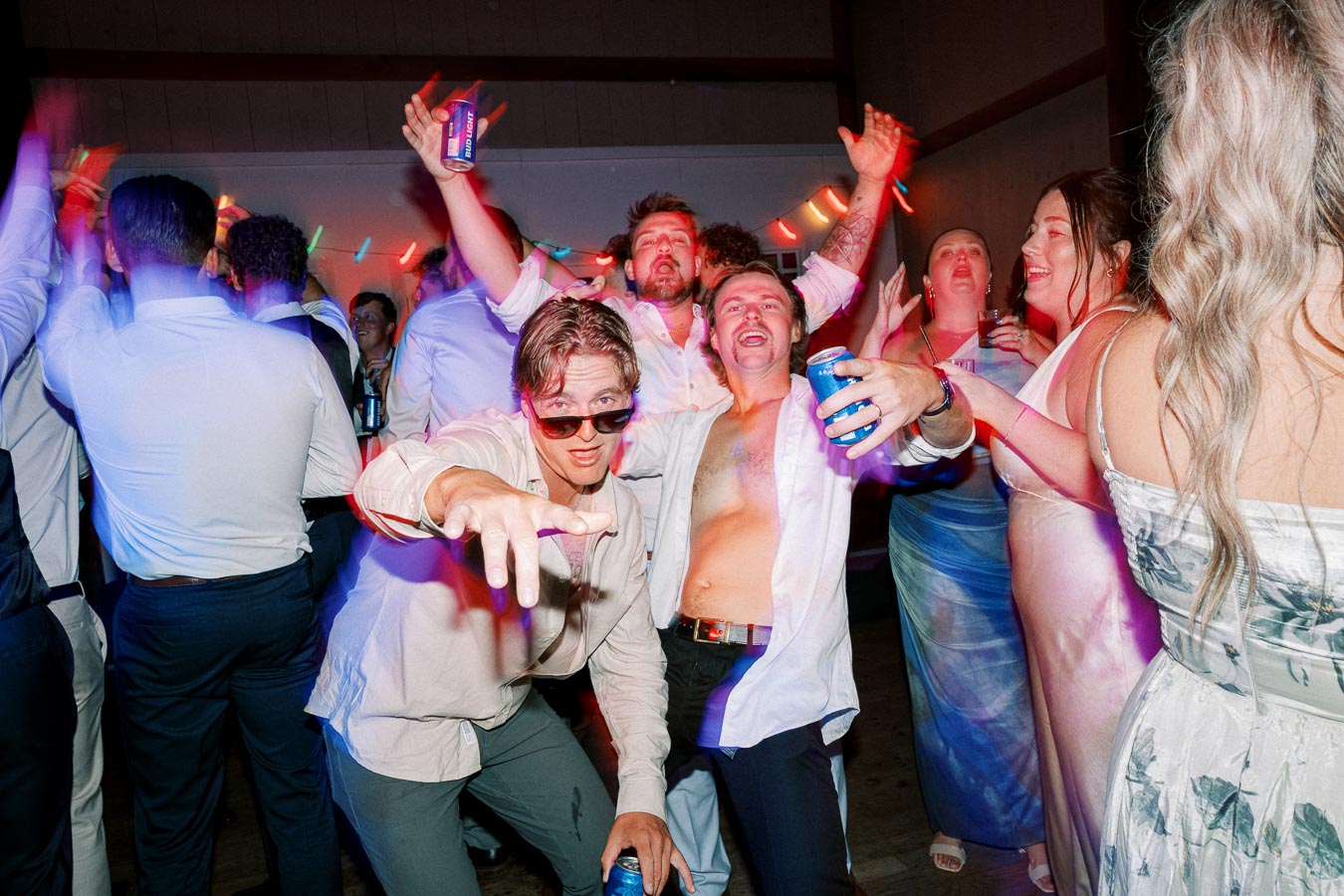 Group of people energetically dancing and having fun at a lively party, holding drinks, with colorful lights adding to the festive atmosphere.